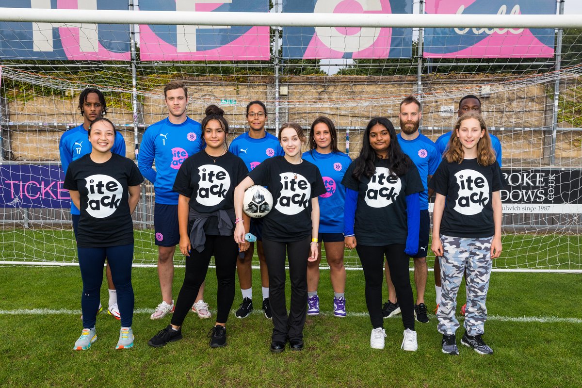 5 young girls in black shirts with a white round Bite Back logo stand in front of 6 footballers clad in blue football jerseys with a pink Bite Back logo.