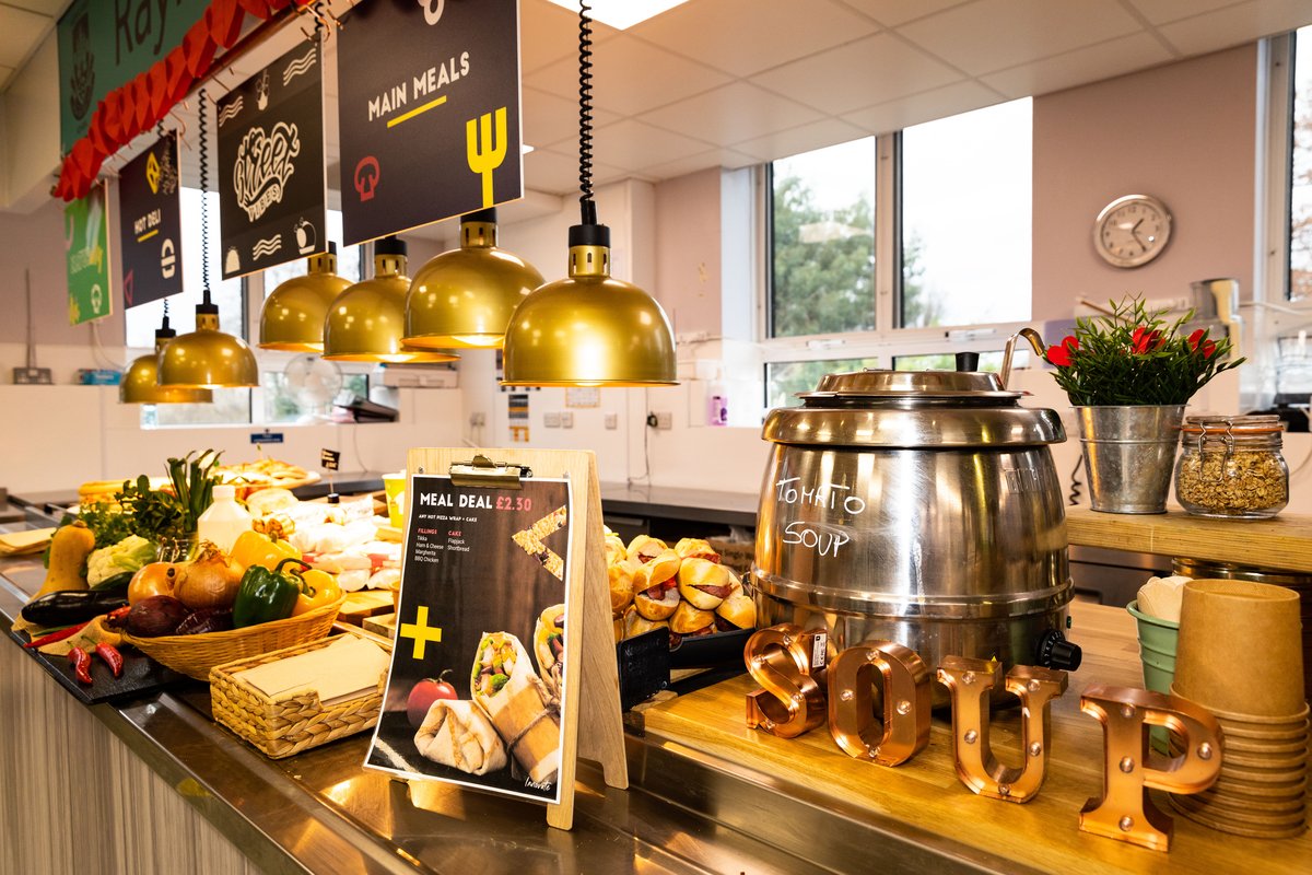 A school canteen counter which has a large soup pot, with 'tomato soup' written on it and in front of the pot, there are gold decorative ornaments spelling out soup. Next to the pot of soup, there is a sign showing meal deals and behind that, a bowl of ho