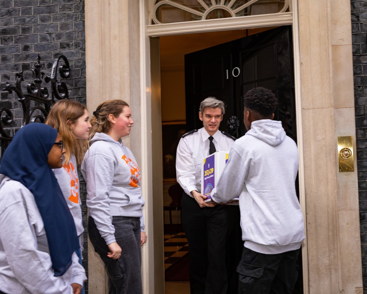 Victor a young black man in a grey sweater is handing over our box with petition signatured to a member of staff at No.10 while three fellow activists in Bite Back sweaters look on.