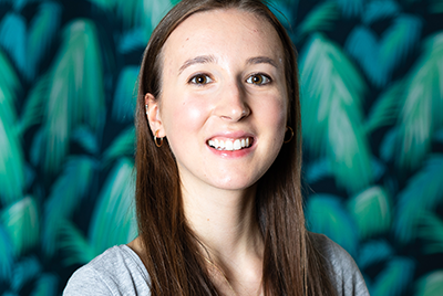 Amy, a young white woman with long, straight brunette hair is smiling at the camera while stood in front of green, leafy wallpaper. She is wearing a grey t-shirt and a necklace with a pendant, and small gold hoops in her ears.