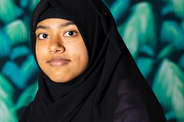 Anica is a young asian girl wearing a black headscarf and is smiling at the camera from a slightly sideways pose. She is stood in front of green, leafy wallpaper.