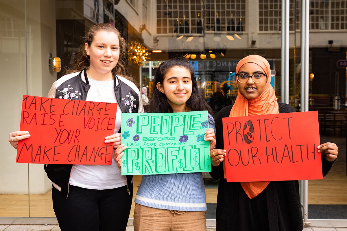 Three teenage girls stand in a row, holding signs reading 'take charge, raise your voice, make change', 'people before fast food company profit' and 'protect our health'.