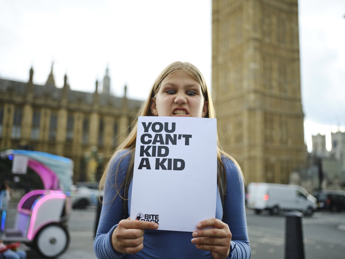 Bite Back activist Maya is standing in front of the Houses of Parliament holding a sign that reads "You can't kid a kid".