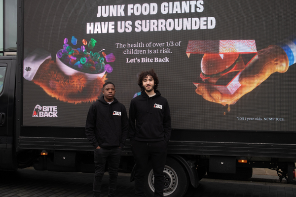 Two young people stood in front of a digivan that displays a billboard that says ‘junk food giants have us surrounded.’ Emmanuel is a young black man with short hair, he is wearing a Bite Back hoodie and black jeans. Reuben is a tall young man with long curly hair and a short beard, he is also wearing a Bite Back hoodie.