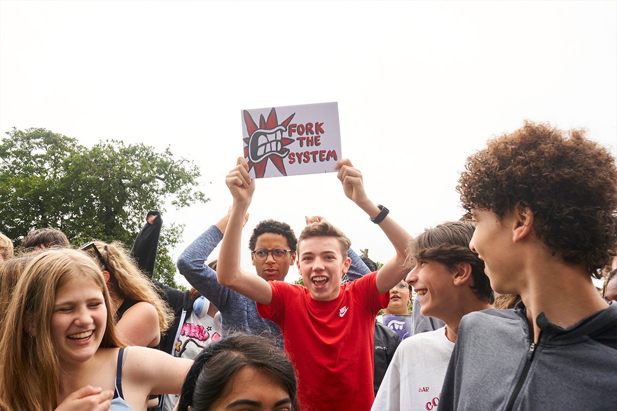 A crowd of young people, with one young man holding a hand-written sign that says 'fork the system'