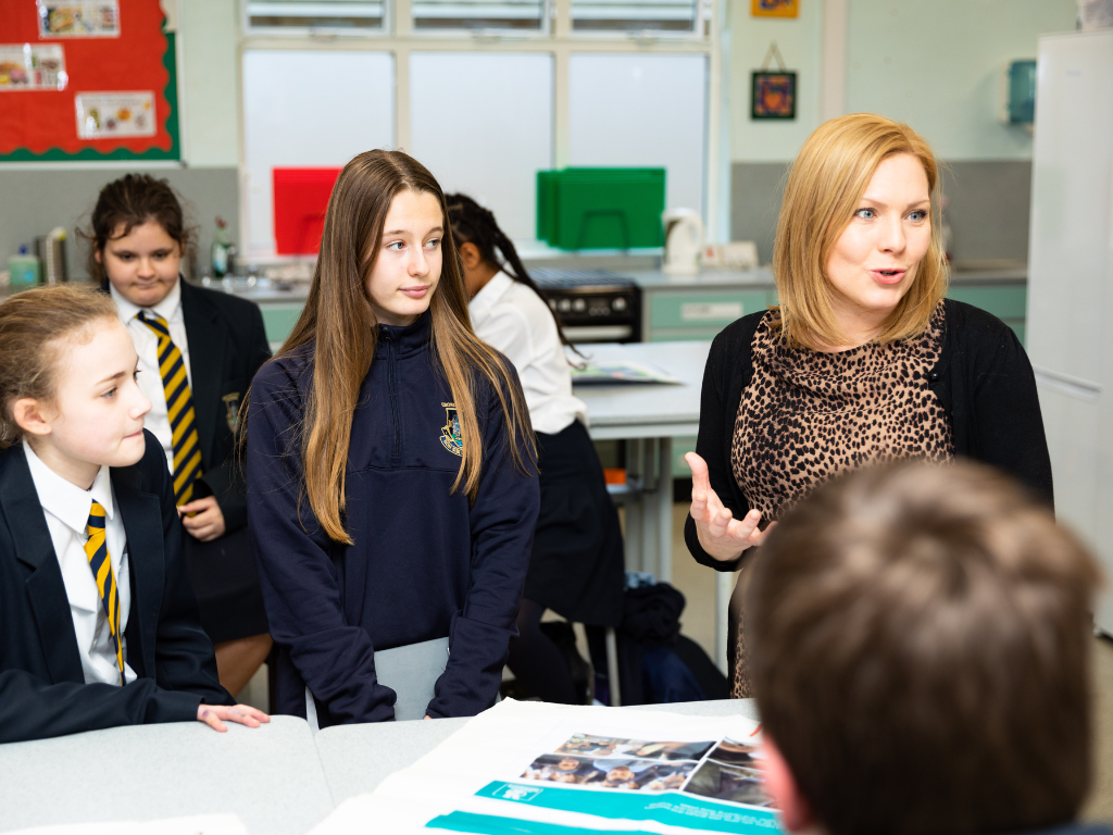 A teacher with a short blonde bob is talking to students, who are dressed in black blazers and striped ties. They are standing around a table and discussing with the teacher in a bright classroom with white walls.