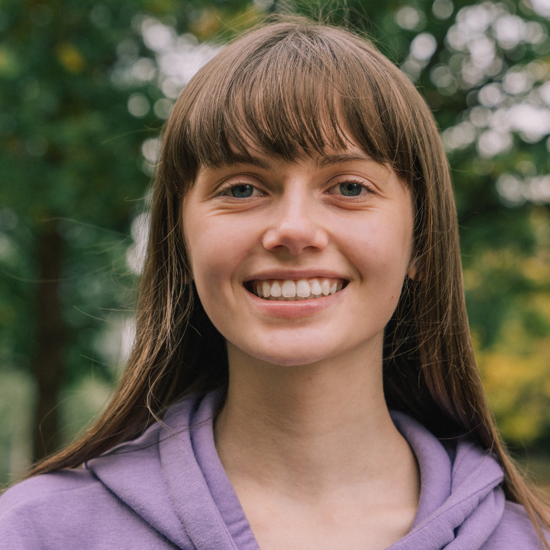 Brooke a young white woman with long brown hair and a fringe is smiling happily at the camera. She is wearing a purple sweater and standing in front of trees.