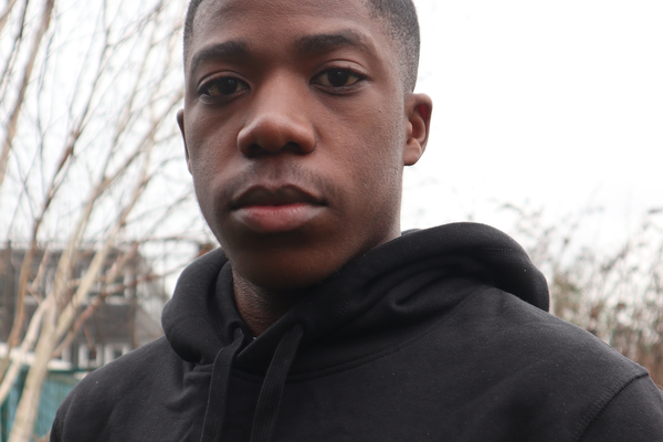 Emmanuel a young black man with short cropped hair and thick lips is looking down into the camera standing in front of a tree lined street. He is wearing a black hoodie.