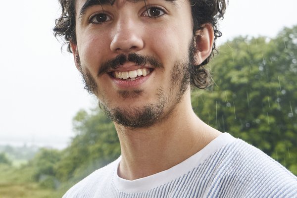 Reuben is a tall tanned man with dark curly hair that falls to his eyebrows and a full beard. He is wearing a white striped t-shirt.