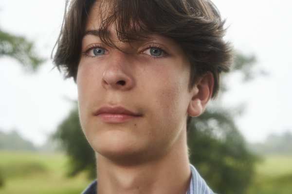 Oli is a young white man with brown messy hair falling down to his ears. He is wearing a black t-shirt with a blue cardigan over it and standing in front of a cloudy sky with trees behind him. He is sporting a serious face showing he is ready to bite back.