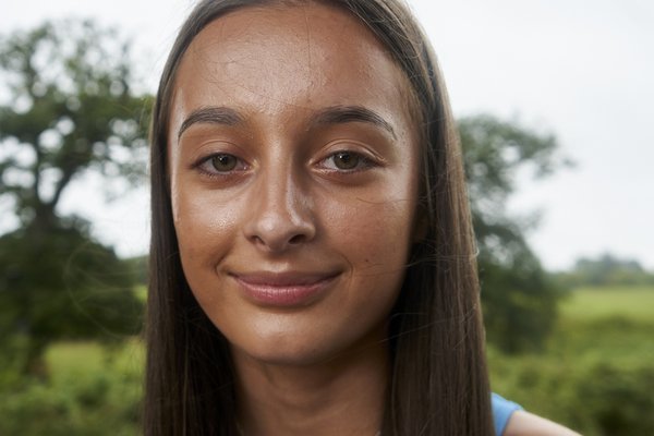 Macey is a tanned young girl with very straight brown hair that hangs below her shoulders. She has a shiny complexion, green eyes, a straight thin nose and a serene expression on her face with the slightest of smiles. Nothing can faze her. She is wearing a blue tank top and a thin silver necklace.