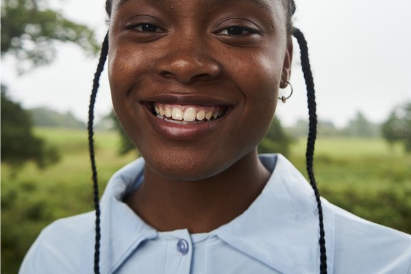 Jayda is a young, black woman with her hair tightly pulled back except for two braids on either side of her face. She is looking into the camera with a powerful expression, and has two small gold hoop earrings in her ears. The blue collar of her shirt can be seen and she stands in front of a field.