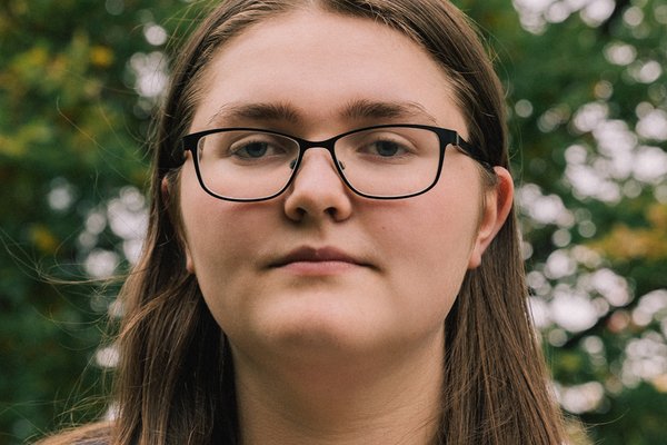 Imogen, a young white person, with long, straight brown hair and wearing thin-framed black glasses, is looking into the camera with a straight face.