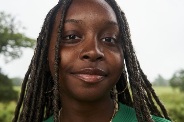 Destiny is a black woman photographed from a powerful angle giving her a strong powerful look. She is a black woman sporting long braids standing in front of a cloudy sky and trees. She is wearing a bright green knitted sweater and a set of thin golden necklaces around her neck.