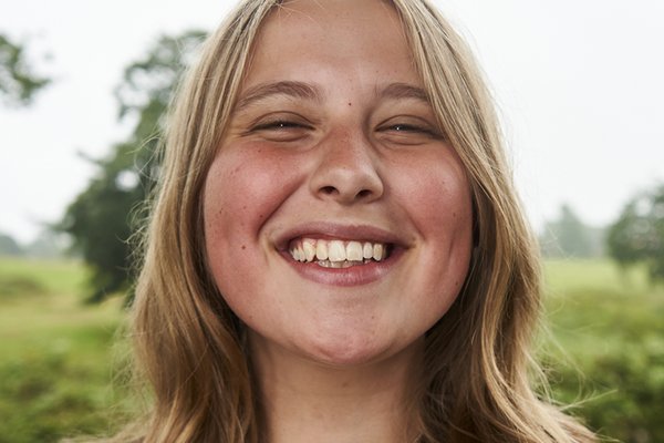 Anna, a young white woman with curly blonde hair, has a big wide grin on her face showing her teeth and scrunching up her eyes and nose. She has rosy cheeks and a stripy jumper on.