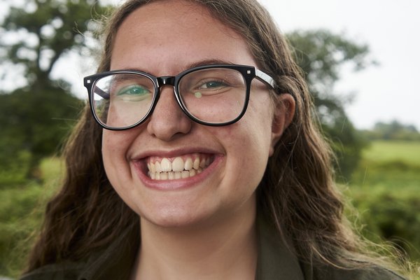 Mia a young white girl with very long curly brown hair wearing black widerimmed glasses and a black suit shirt. She is standing in front of a cloudy sky with green trees behind her. She is sporting a big smile to show she is ready to tackle a system set up to fail us all.