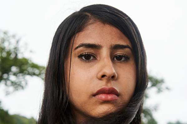 Nabeeha, a young brown-skinned woman, with straight, jet-black hair is looking into the camera from a side angle with a serious expression on her face. She is wearing light makeup.