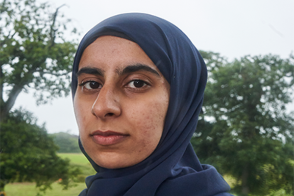 An image of Faheemah in front of a sky with green trees behind her, she's a brown skinned girl wearing a dark blue hijab and sporting a smile showing she is ready to take a bite out of the food system