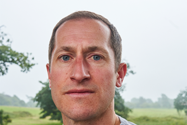 James is a white-skinned man with short brown hair and a light blue top. He is staring into the camera with a serious expression showing he is ready to bite back, and behind him, you can see the trees in the scenery.