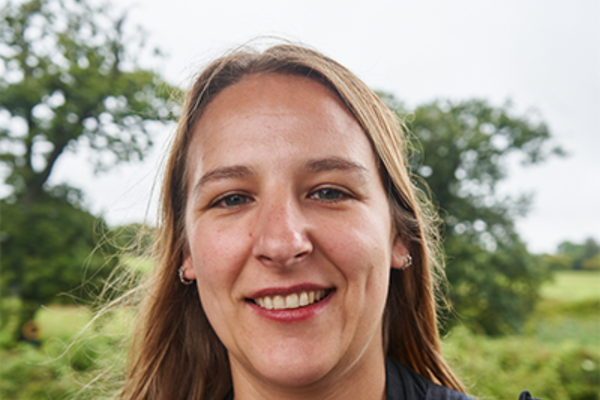 Rachel has long blonde hair and is smiling into the camera with a big smile. She is wearing a black tshirt and small hoop earrings and behind her, there are trees and a field. Her hair is flowing in the wind.