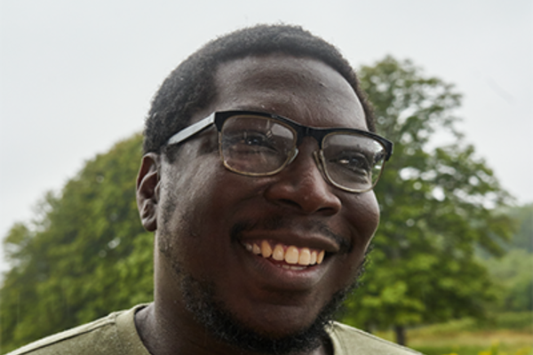 Aaron is a black man with short hair and black, rectangular framed glasses. He is facing the camera from a sideways angle with a smiley expression ready to bite back! Behind Aaron, the background is full of trees and greenery