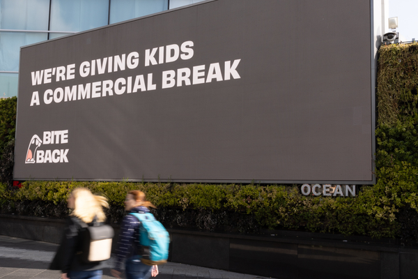 A large black billboard with white writing saying "We're giving kids a commercial break." displaying at Westfield White City with two people passing by in a blur.