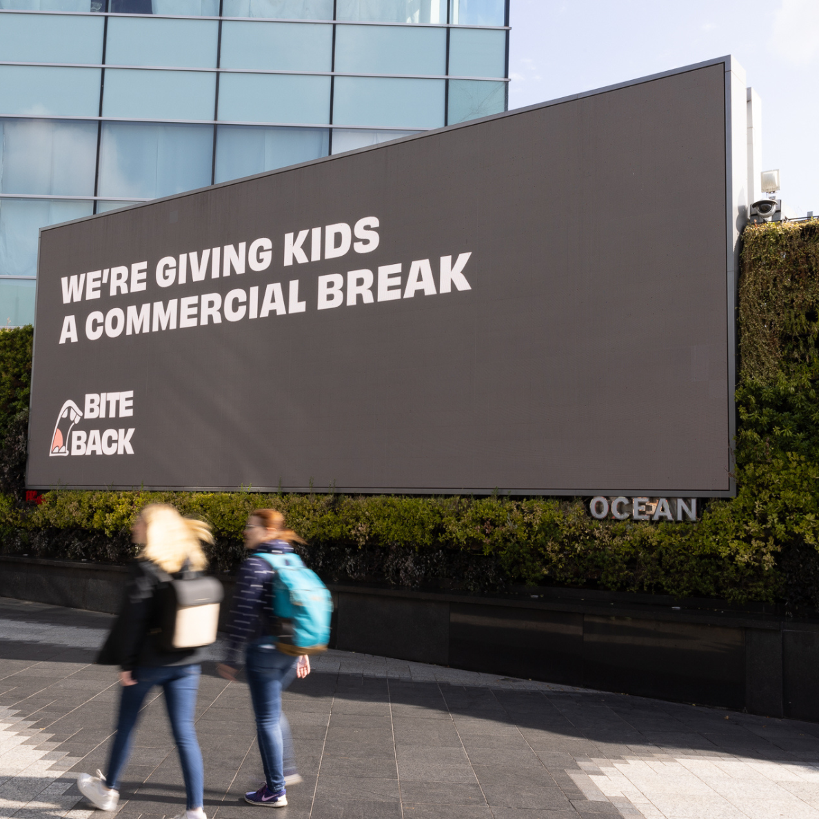 A large black billboard with white writing saying "We're giving kids a commercial break." displaying at Westfield White City with two people passing by in a blur.