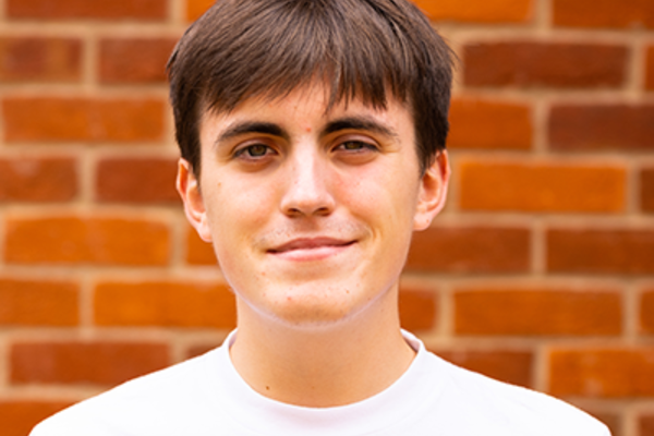 James, a young white man with floppy brown hair falling on his forehead, is smiling at the camera and stood in front of a brick wall.