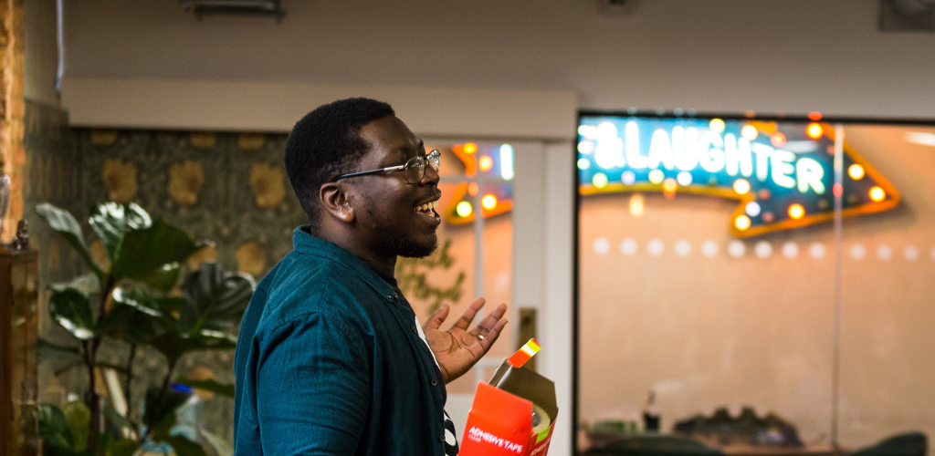 Aaron is a black man with short hair and a green shirt. He is speaking to a group of people in the JO office and gesturing towards them, holding a box in his right hand. He wears rectangle framed glasses.
