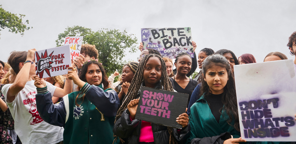 A group of young people stood in a field holding protest signs up. At the front of the group, there is a young black woman with blonde and black braids who is wearing a black jacket and holding a sign painted in black with purple lettering that reads 'Show Your Teeth' in all caps