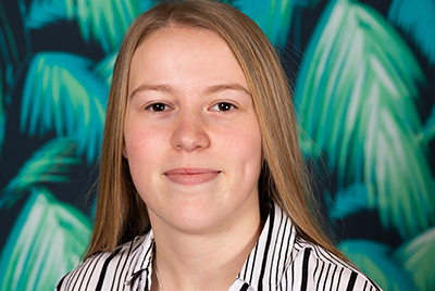 Lily is wearing a pinstriped shirt and is smiling at the camera. Her straight blonde hair is falling down her back and rests lightly on her shoulders.