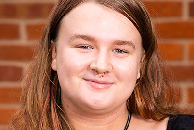 A young blonde woman is stood in front of a wall. Her hair is neatly parted and she is smiling at the camera