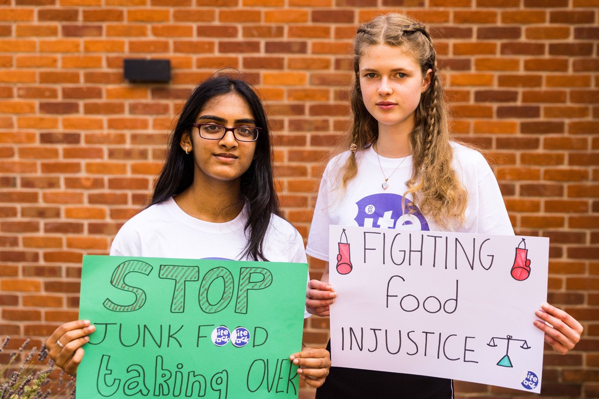 Two teenage girls stand in front of a brick wall, holding handwritten signs reading 'stop junk food taking over' and 'end food injustice'.