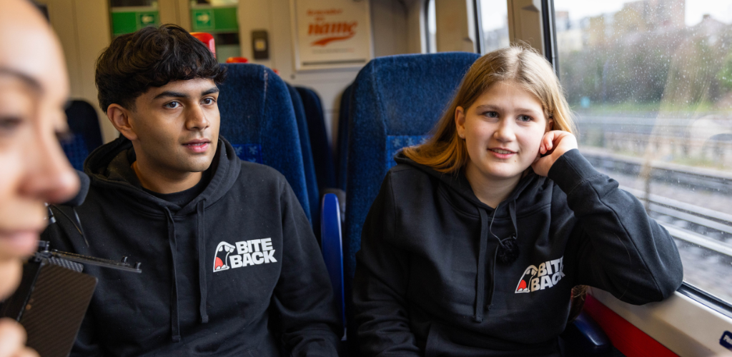 Dev is a young brown skinned man with a black Bite Back hoodie on. He is sitting on a train with Maya, a young blonde woman who is also wearing a black Bite Back hoodie. They are speaking to the cameraman sitting opposite them.