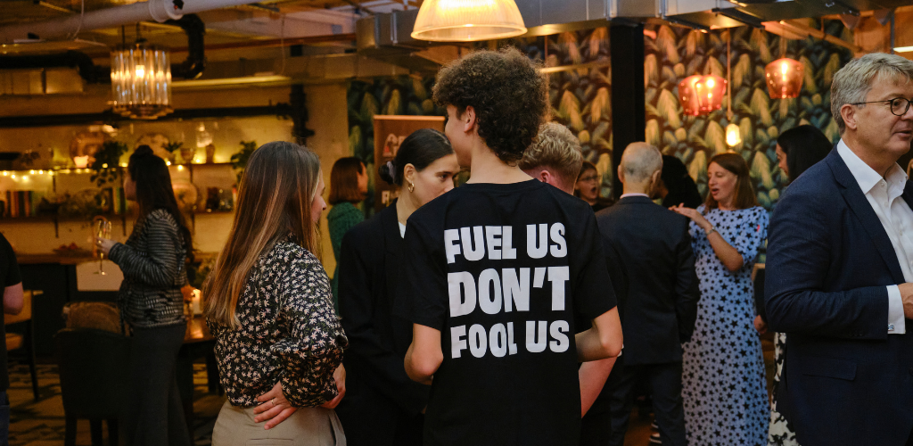 A young person with brown curly hair has their back to the camera. They are wearing a tshirt that reads 'Fuel Us, Don't Fool Us' in large writing at the back