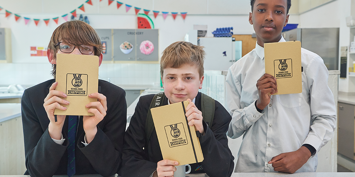 Three School Food Champions one white boy with long floppy brown hair, one with  short blond hair and a taller black boy all holding up neige notebooks with the School Food Champions logo