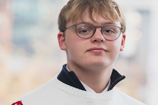 Sam is a young white man with cropped blond hair. He wears thin-framed glasses and a red and white patterned jumper. The collar of his shirt is visible from the top of the jumper. He is stood in front of a large window.