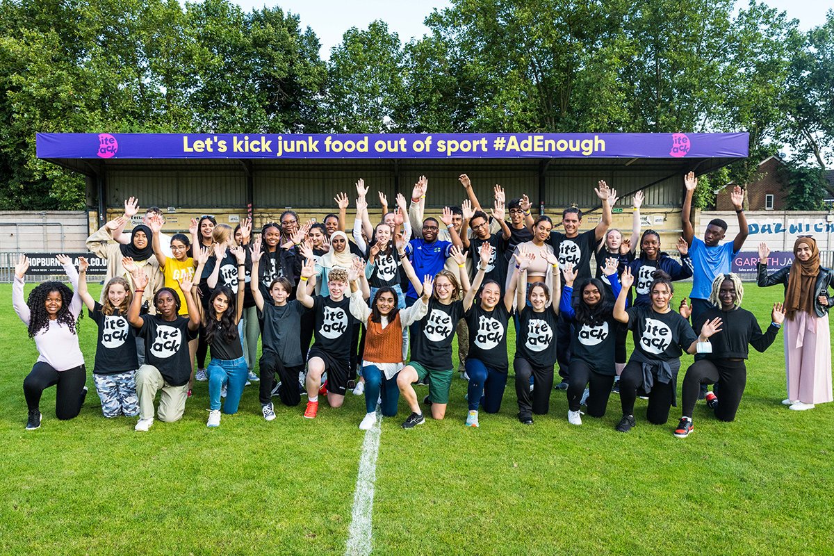 A crowd of teenagers stand in Dulwich Hamlet FC stadium in front of a sign saying 'let's kick junk food out of sports'.
