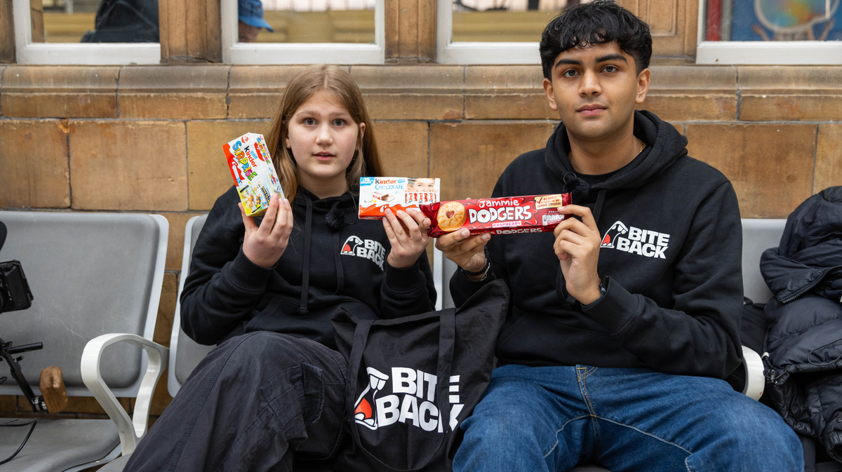 A young blonde woman and a brown skinned man with cropped black hair are sat on a bench in a train station. They are wearing black Bite Back hoodies and are holding up unhealthy products with child-appealing marketing on them.