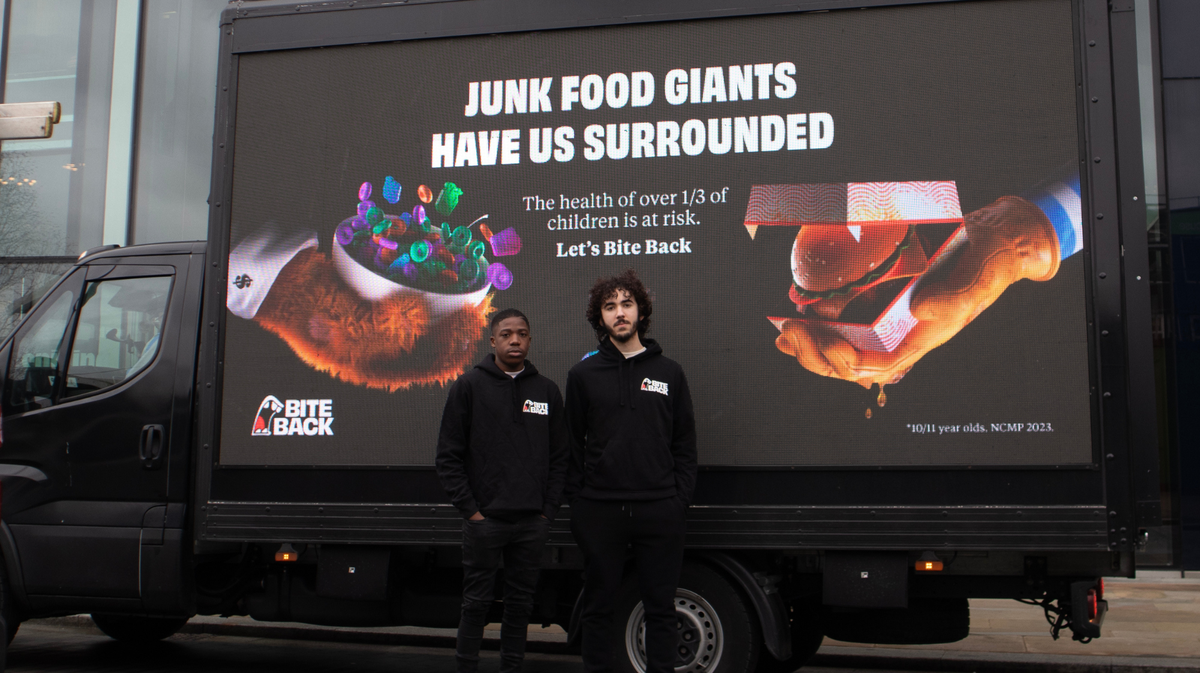 Two young men are stood in front of a Digi Van that displays a poster saying 'junk food has us surrounded'.