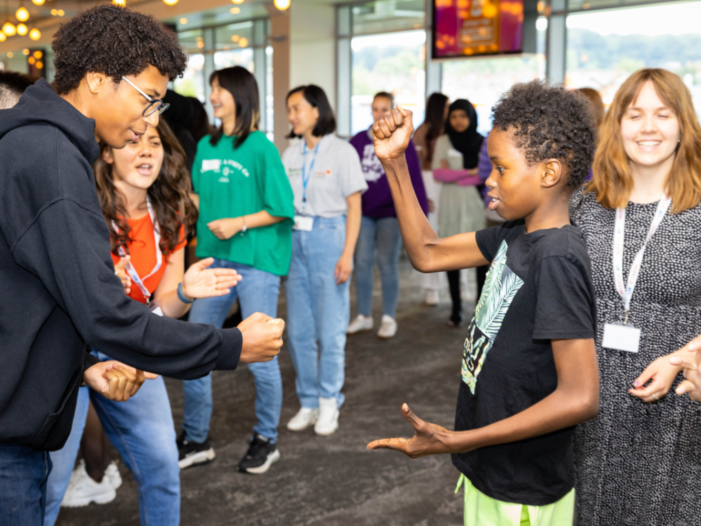 Alex, a young black male with short curly hair and glasses, is playing rock paper scissors with a younger black boy. Alex is wearing a navy hoodie and the young boy is wearing a black graphic t-shirt and green shorts.