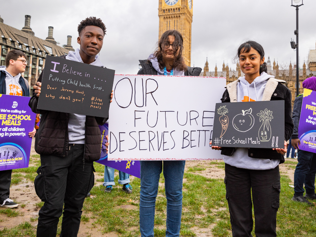 Victor standing together with two fellow activists holding a protest sign in front of Big Ben. He is a black young man with short cropped hair, wearing a black jacker and gray T-shirt holding a sign that reads "I believe in...putting Child Health First."
