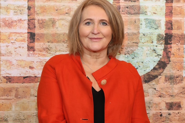 Anne is a white woman with a blonde bob cut. She is wearing a red coat with a black top underneath and is stood in front of a brick wall and smiling at the camera.
