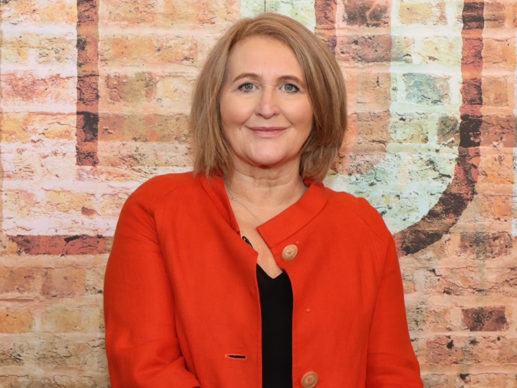 Anne is a white woman with a blonde bob cut. She is wearing a red coat with a black top underneath and is stood in front of a brick wall and smiling at the camera.