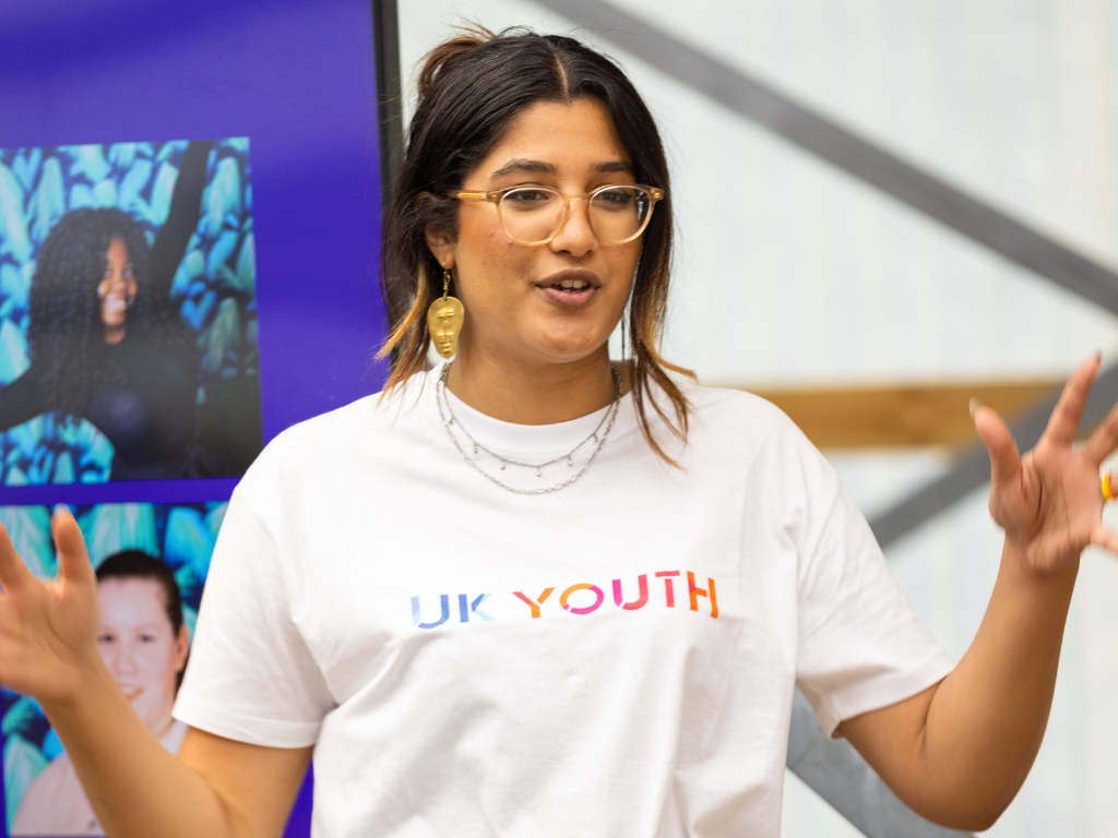 A brown-skinned woman, with long dark hair tied in a bun and large, gold glasses, is speaking to a crowd of young people (not pictured). She is gesturing with her hands in the air and is wearing a white t-shirt that says 'UK Youth' on it and a necklace that hangs over her tshirt.