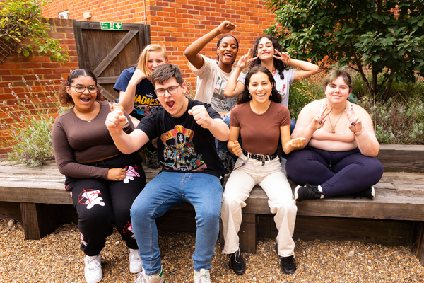 A group of young people sat outside on a bench, with greenery in the background and gravel on the floor. They are holding their arms in the air and smiling, with victorious expressions on their faces.