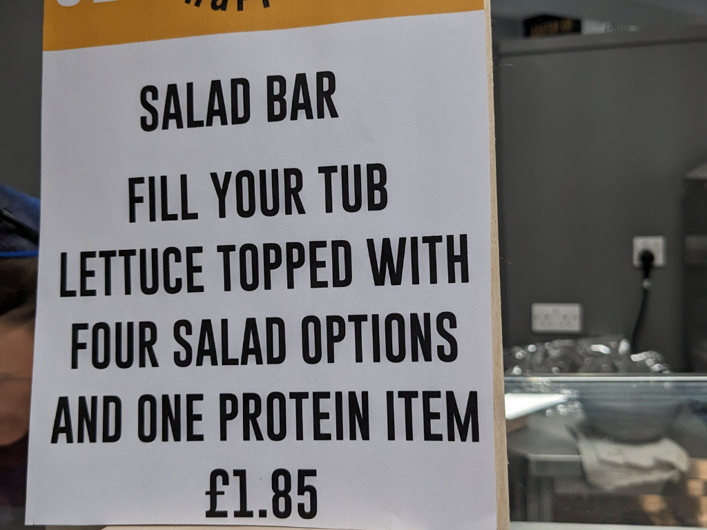 A sign for a salad bar listing prices. The text reads, 'Fill your tub, lettuce topped with four salad options and one protein option. £1.85'