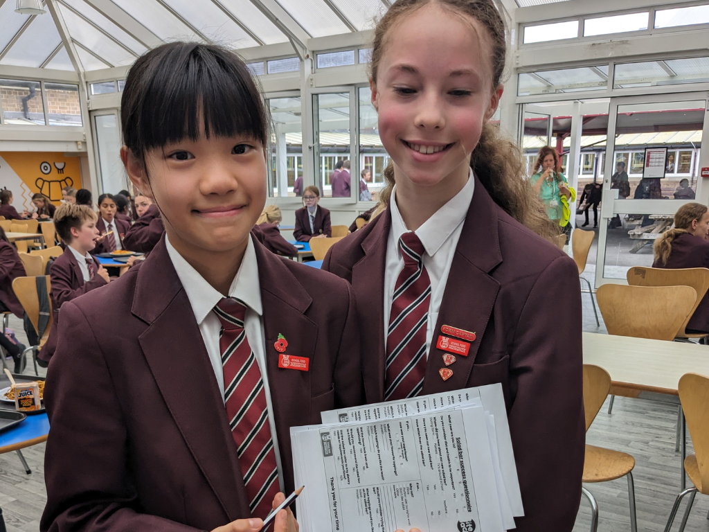 Two young girls, in black and maroon school uniforms, are smiling at the camera while holding a pile of survey sheets and pens in a school canteen.