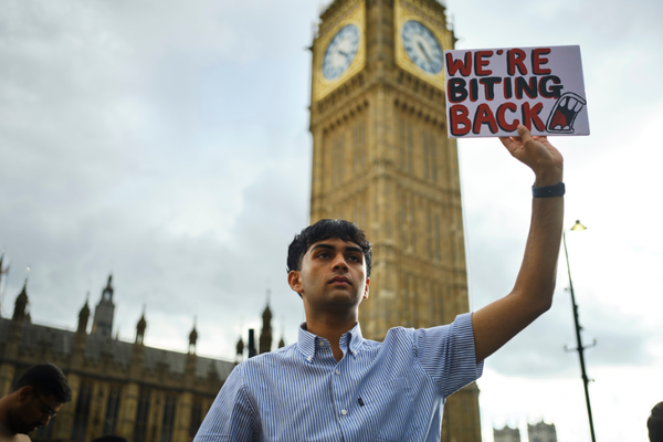 Dev is a young brown-skinned man with short black hair and a light blue shirt on. He is stood in front of Westminster with a serious expression on his face and is holding up a protest sign that reads ‘we’re biting back’
