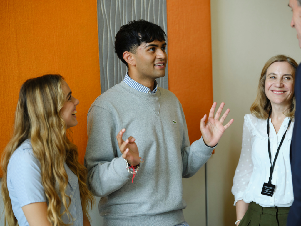 Dev is a young brown-skinned man with short black hair and a grey jumper on. He is stood in a room addressing a member of Parliament, and has his hands gesturing as he speaks. He is stood in between Alice, a young blonde girl, and Caroline, a woman with reddish-brown hair.